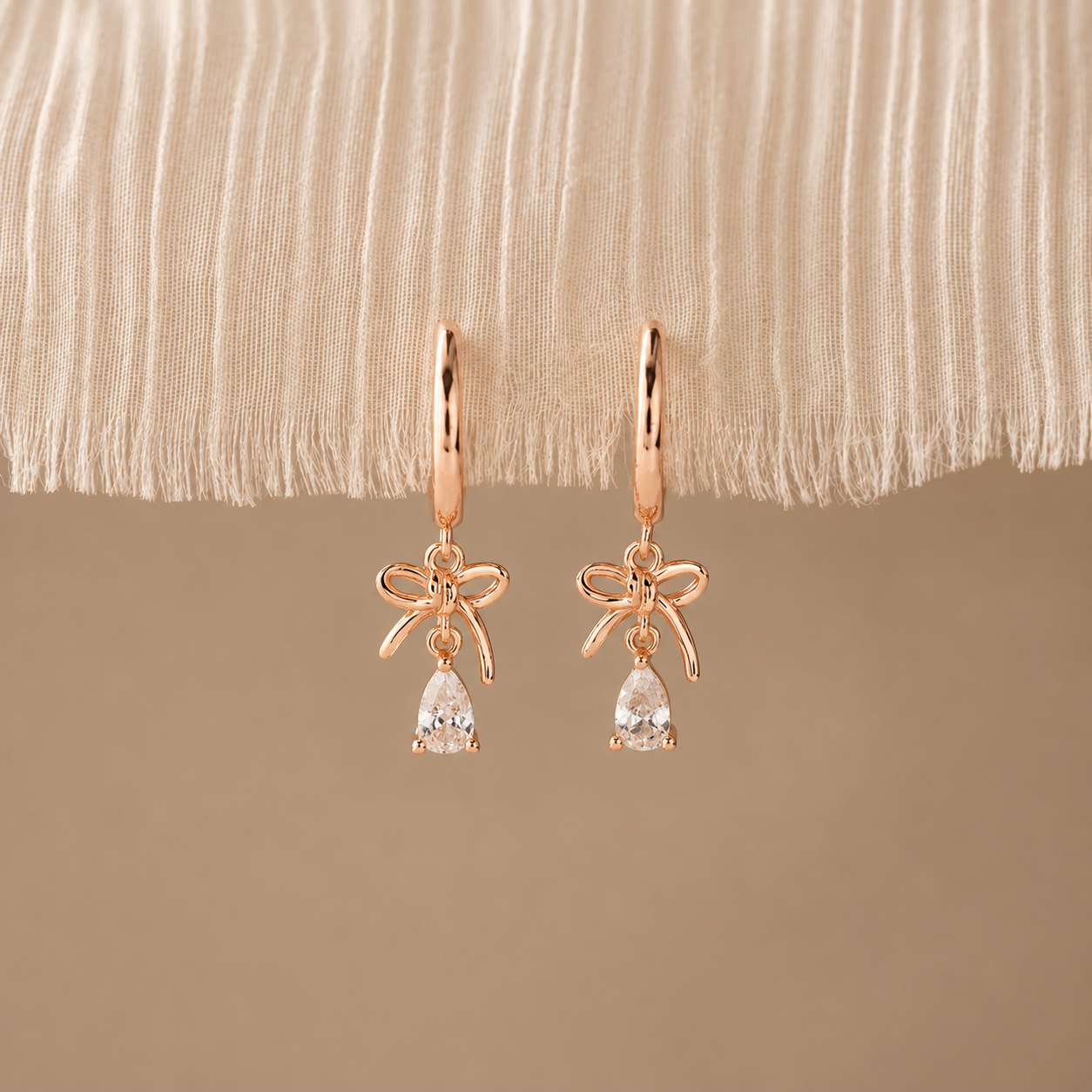 Rose gold bow-shaped earrings with clear gemstones on a beige fabric background