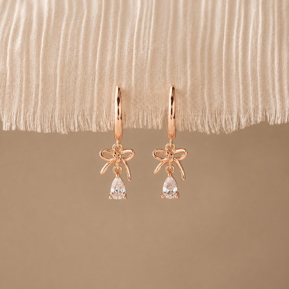 Rose gold bow-shaped earrings with clear gemstones on a beige fabric background