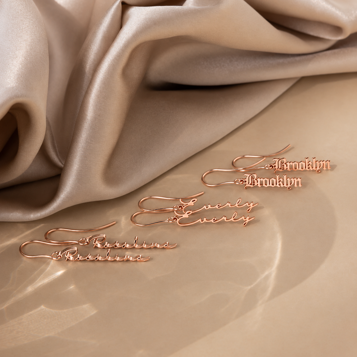 Rose gold earrings with 'Brooklyn' and 'Evelyn' inscriptions on a beige fabric background