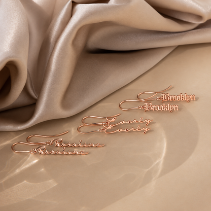 Rose gold earrings with 'Brooklyn' and 'Evelyn' inscriptions on a beige fabric background