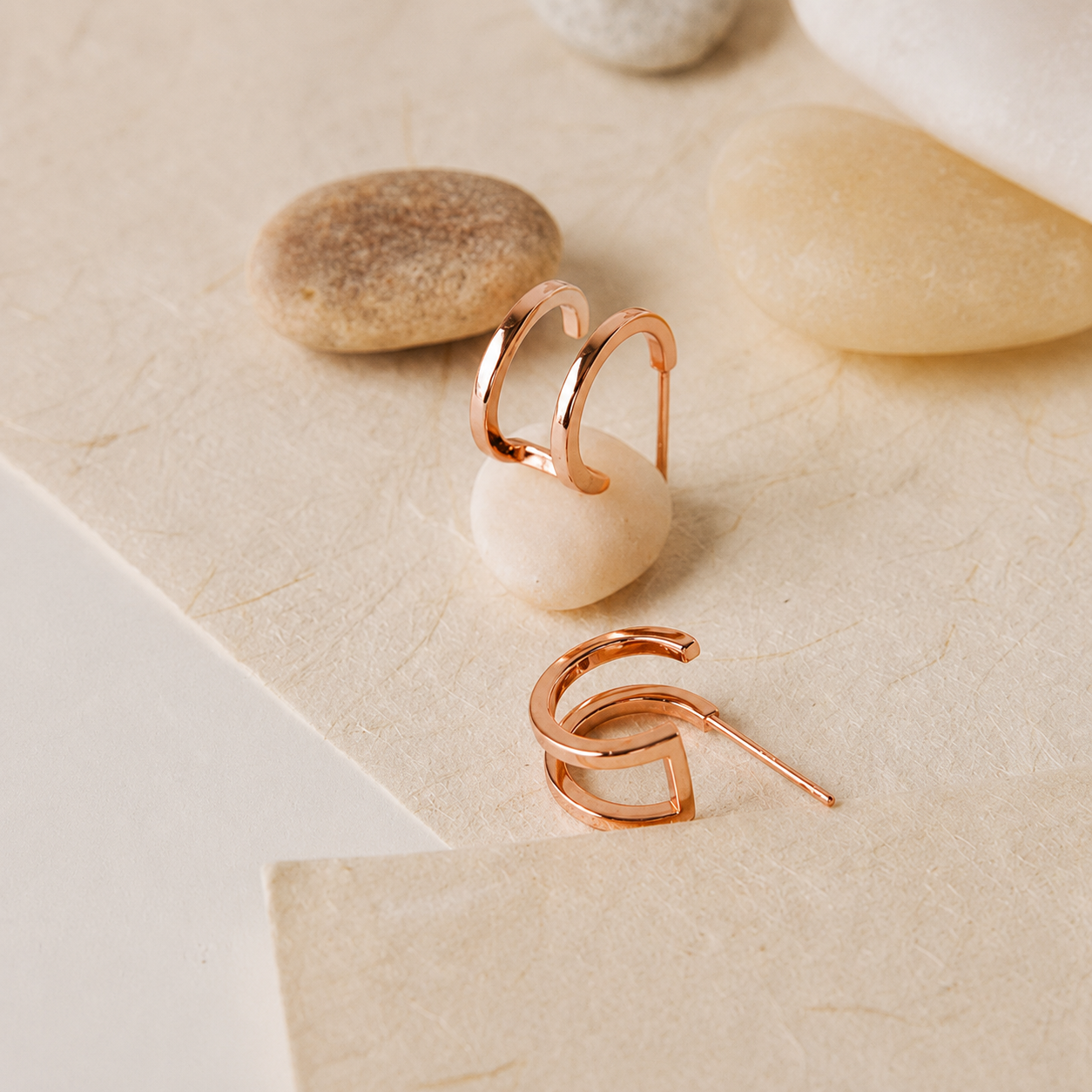 A pair of open hoop earrings in polished rose gold, styled on a textured beige surface with smooth stones.