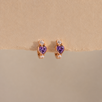 Heart-shaped purple gemstone earrings with rose gold setting on a beige background