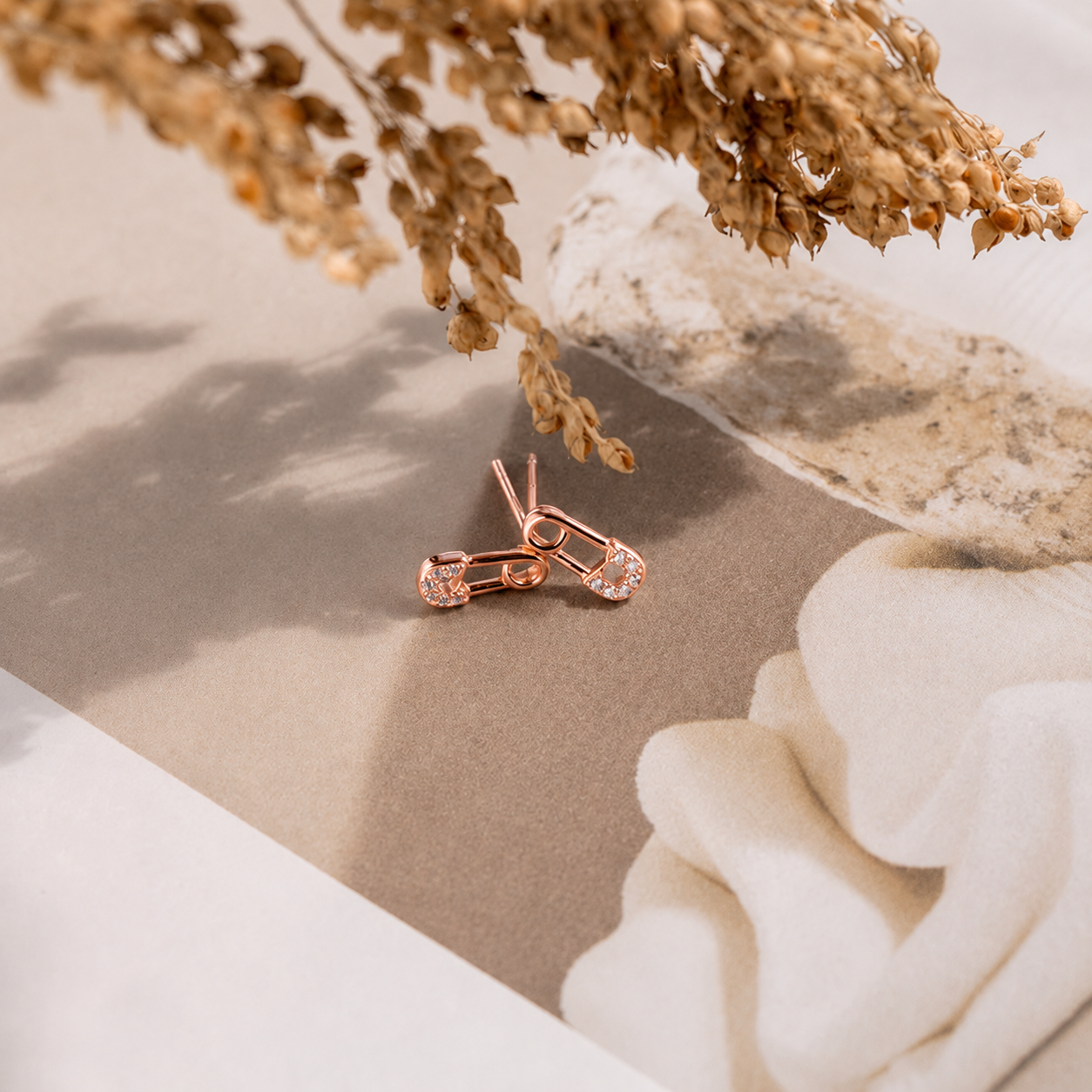 Rose gold earrings on a neutral surface with dried flowers and a white flower.