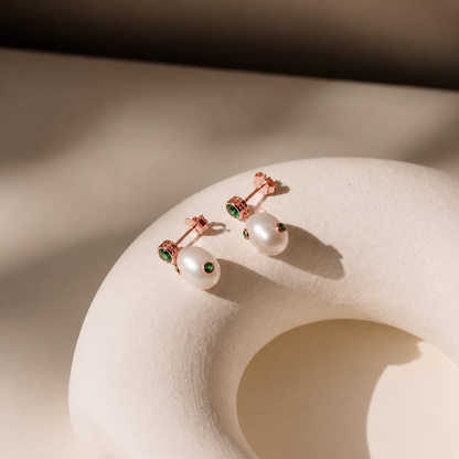 Pair of pearl earrings with rose gold and green gemstone accents on a beige stone surface.