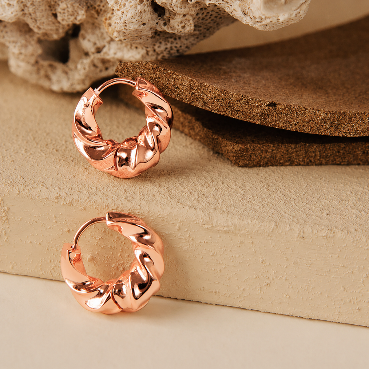 A pair of rose gold hoop earrings with a textured, twisted swirl design, displayed on a beige stone surface with cork and a dried flower prop.
