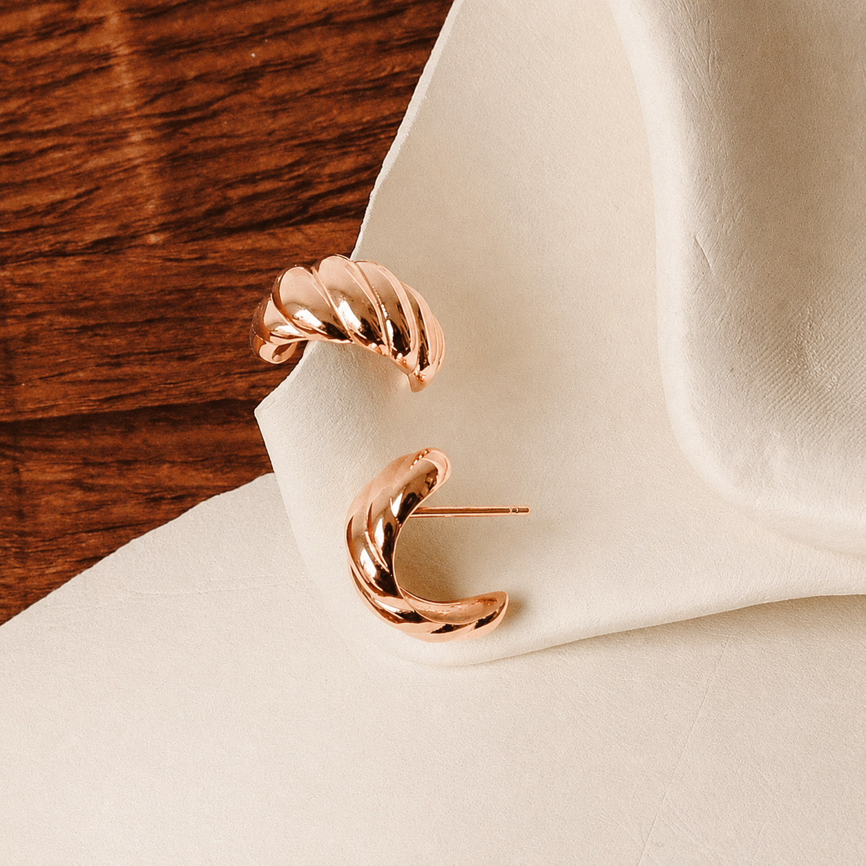 A pair of twisted crescent-shaped rose gold earrings displayed on a draped cream fabric surface next to a dark wood background.