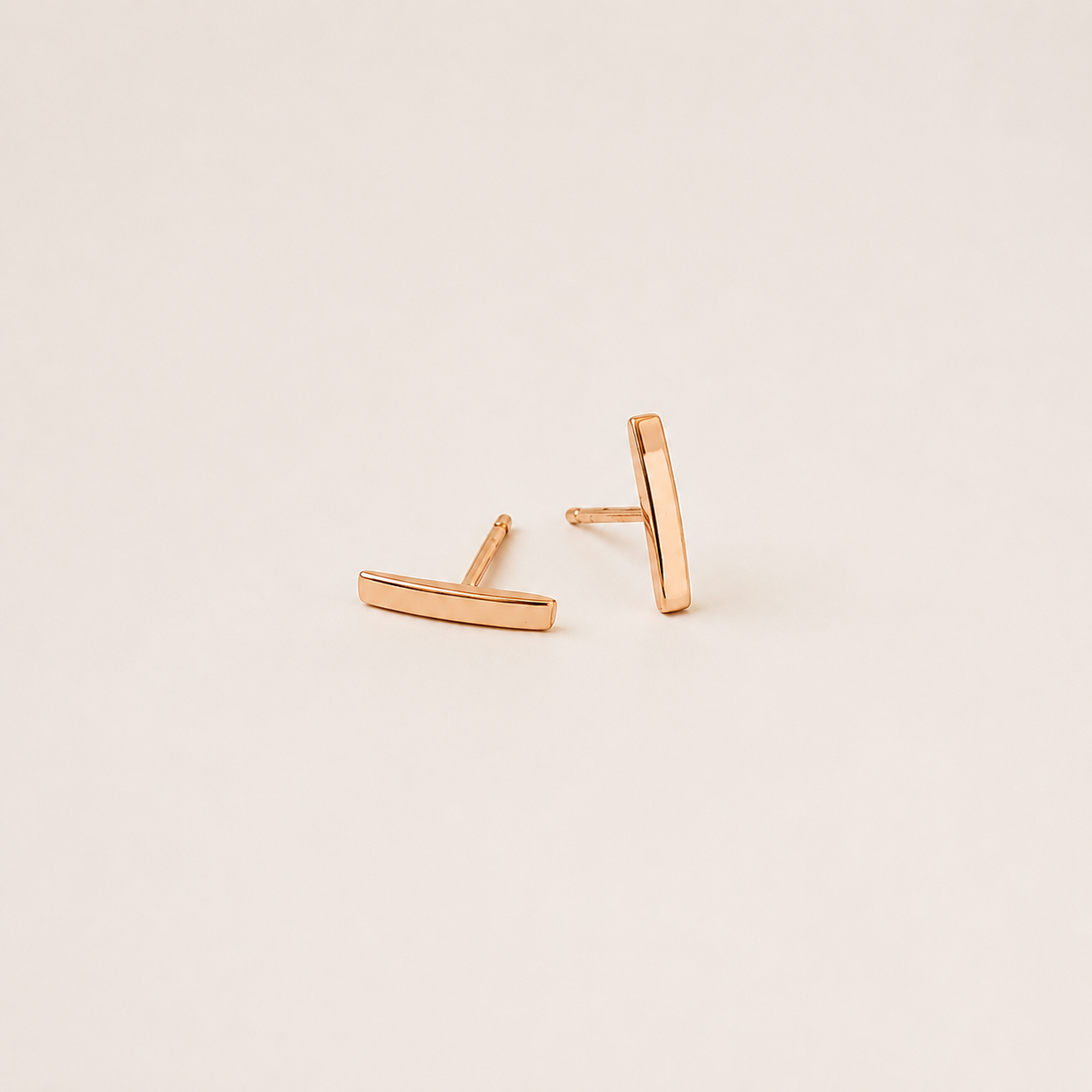 A pair of minimalist rose gold geometric stud earrings, one shaped like a horizontal bar and the other like a vertical bar, displayed on a clean, solid, warm off-white background.
