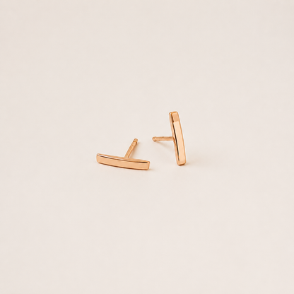 A pair of minimalist rose gold geometric stud earrings, one shaped like a horizontal bar and the other like a vertical bar, displayed on a clean, solid, warm off-white background.