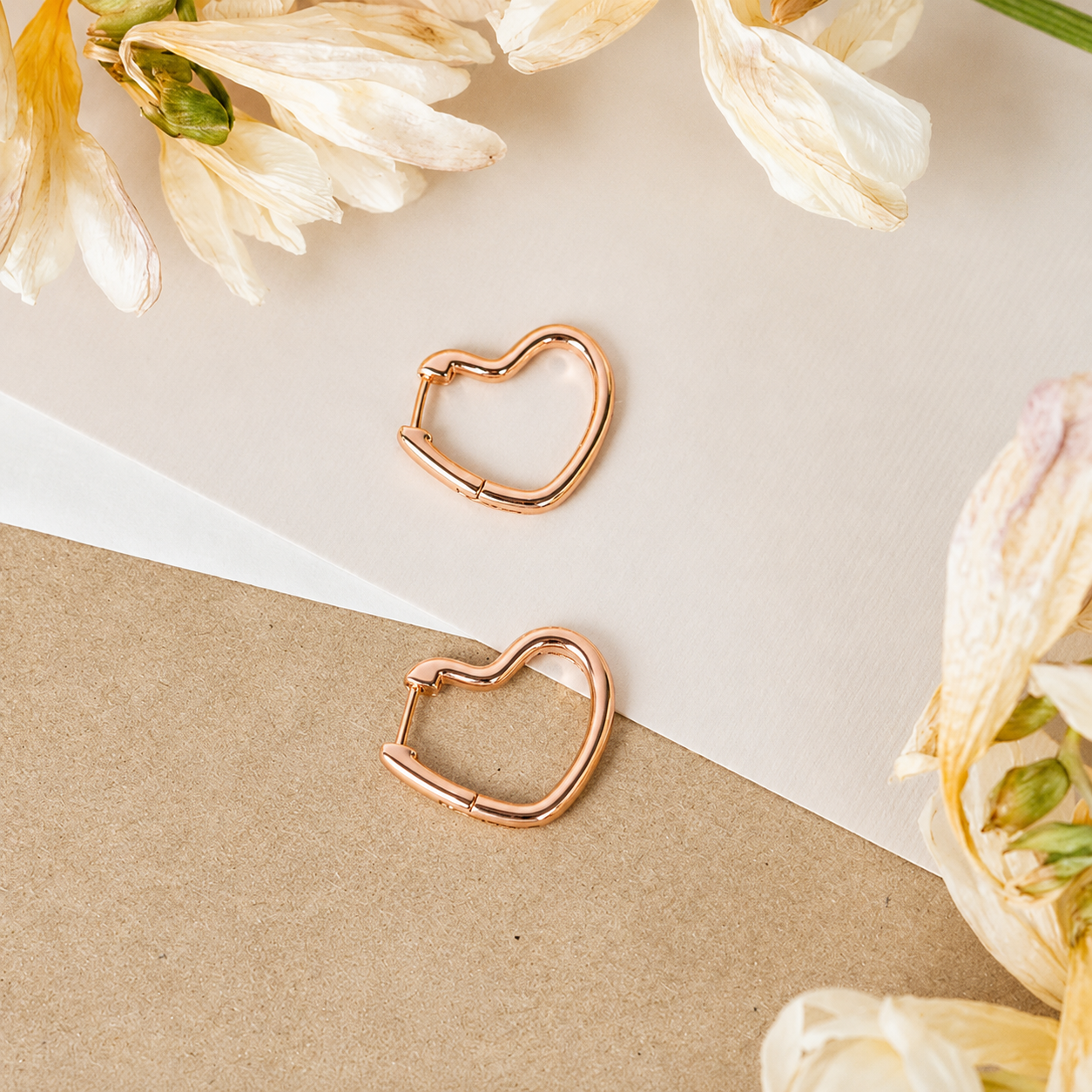 Rose gold heart-shaped hoop earrings on a beige surface with white flowers.