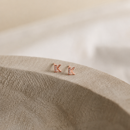 Rose gold 'K' letter earrings on a textured beige surface with a soft background