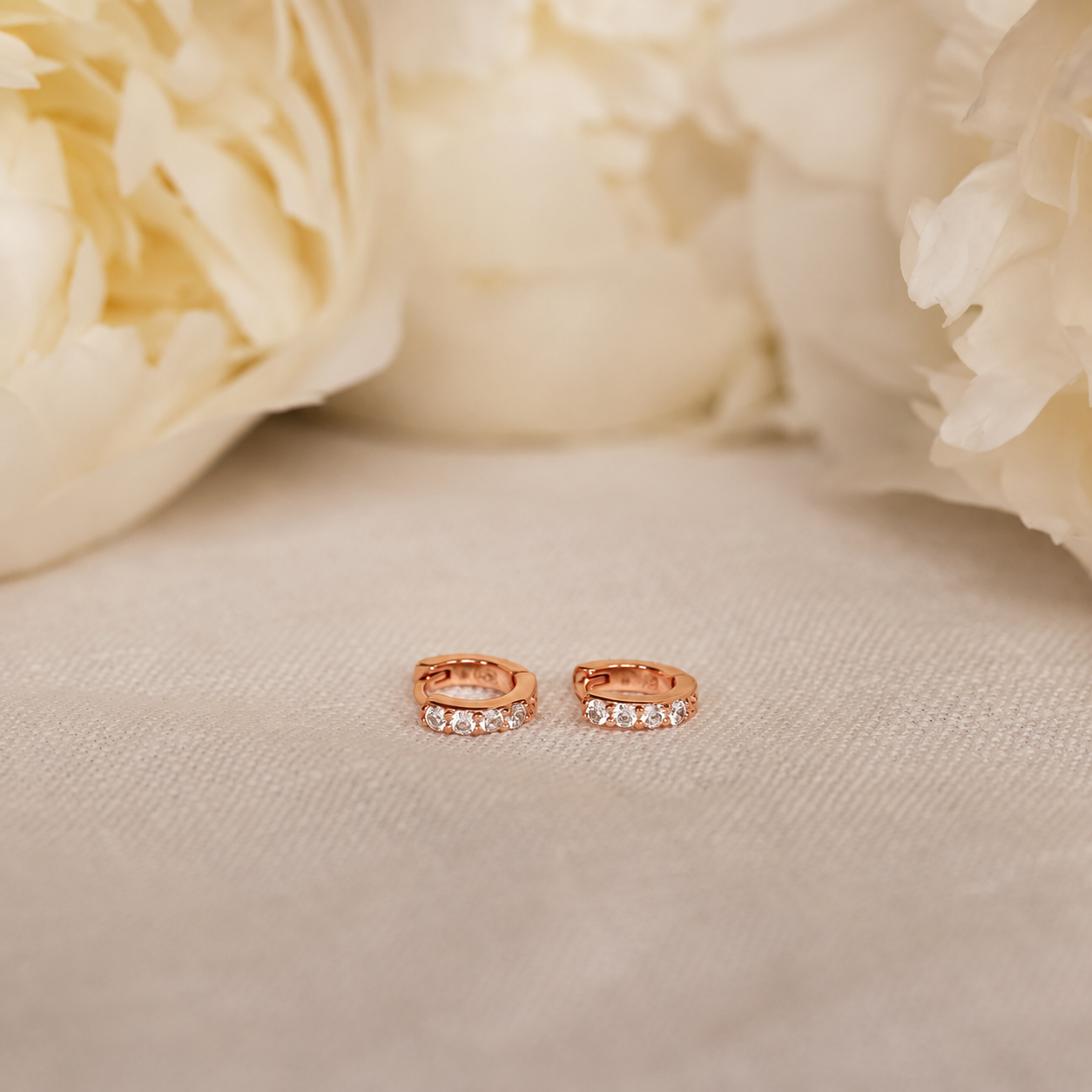 Rose gold hoop earrings with small diamonds on a beige fabric background with cream flowers.