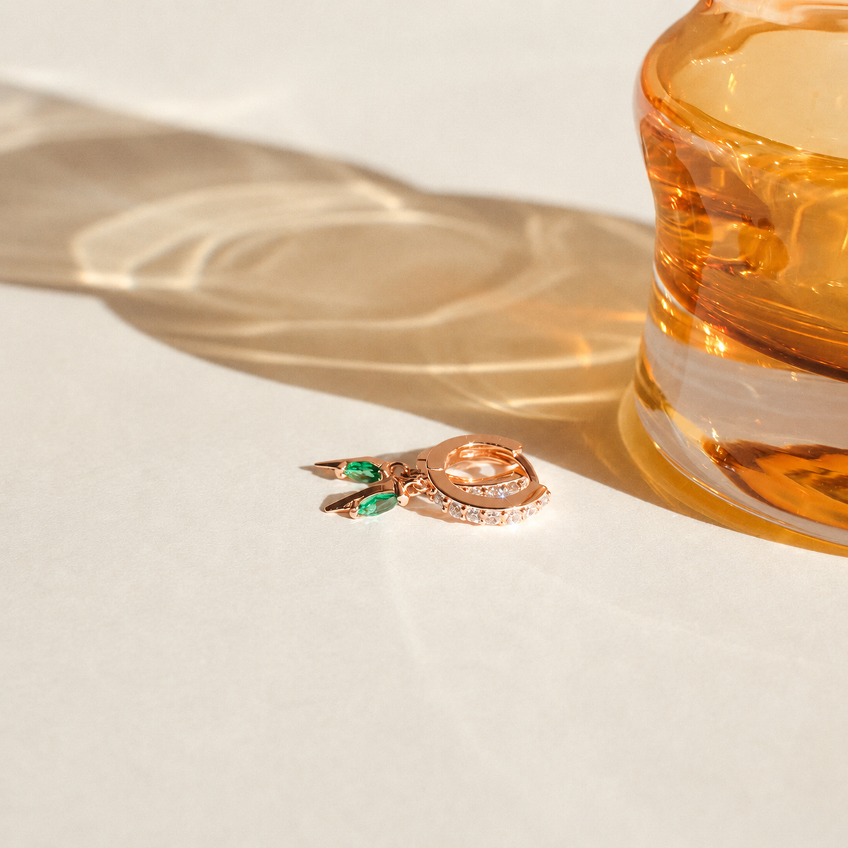 Two gold rings with green gemstones on a light surface next to an orange glass container.