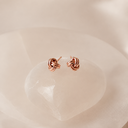 A pair of small rose gold knot stud earrings resting on a smooth, pale pink stone against a soft, warm beige background.