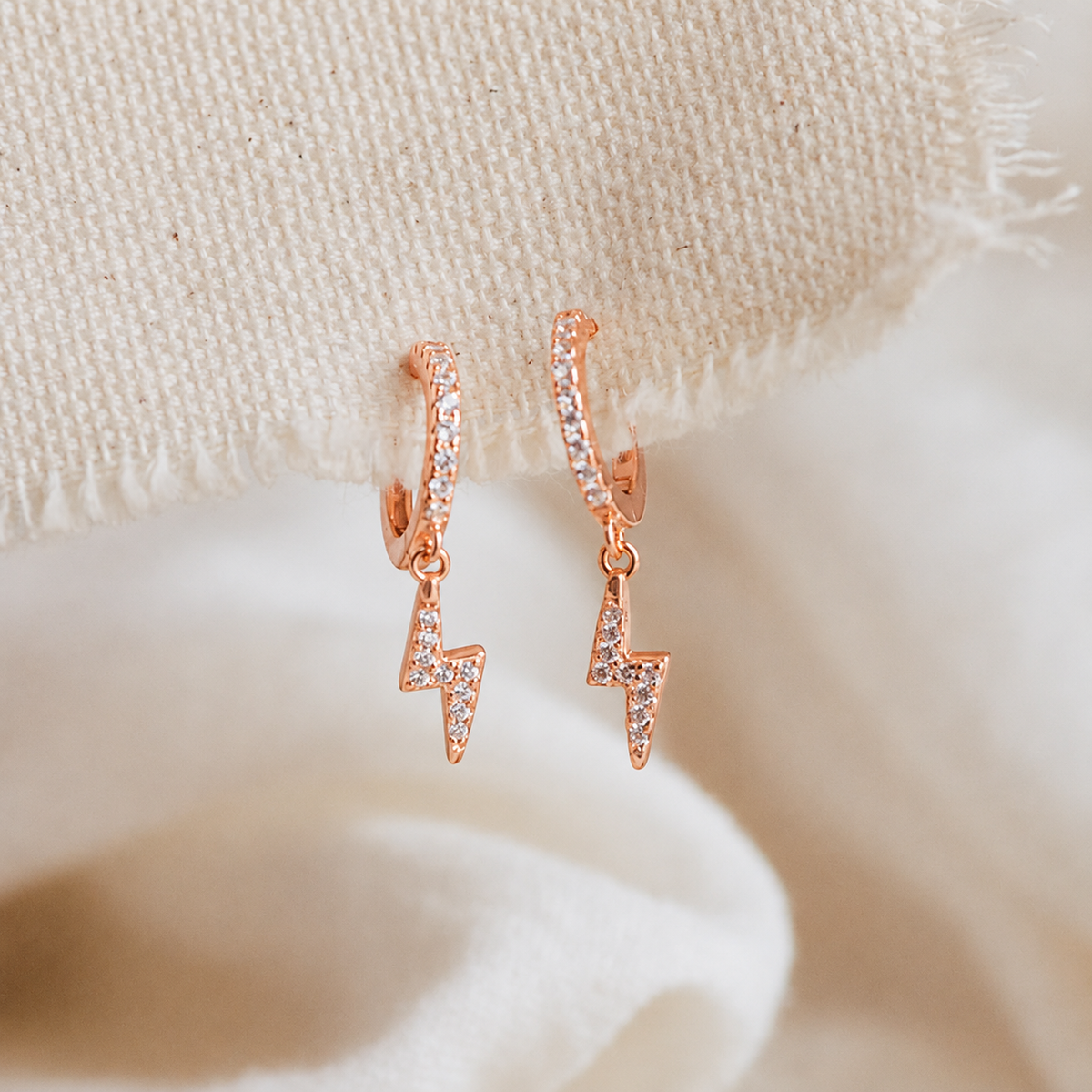 Rose gold lightning bolt earrings on a soft beige fabric background