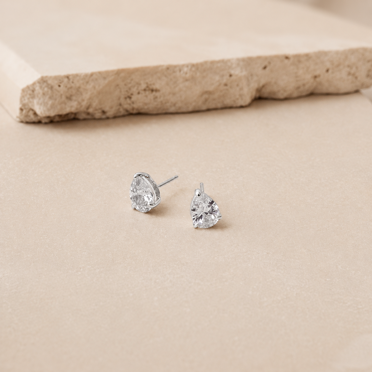 A pair of silver stud earrings with heart-shaped clear gemstones, displayed on a beige textured surface with a stone prop in the background.