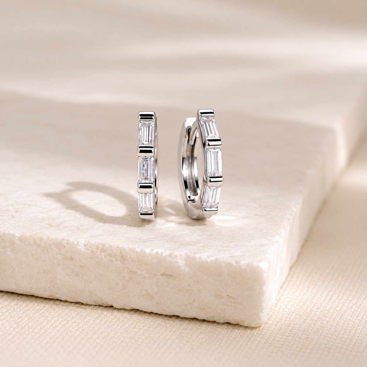 A pair of silver hoop earrings with baguette-cut clear stones, displayed on a textured cream stone slab.