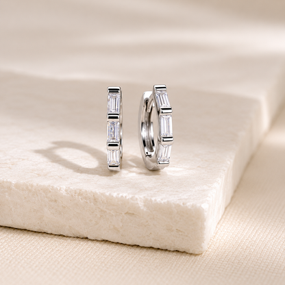 A pair of silver hoop earrings with baguette-cut clear stones, displayed on a textured cream stone slab.