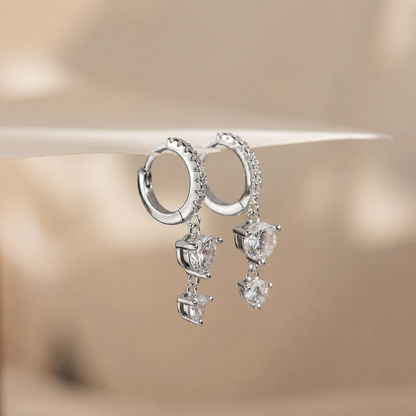 A pair of silver hoop earrings with small clear stones along the top of the hoops and three dangling clear heart-shaped and round stones, displayed against a soft beige background.