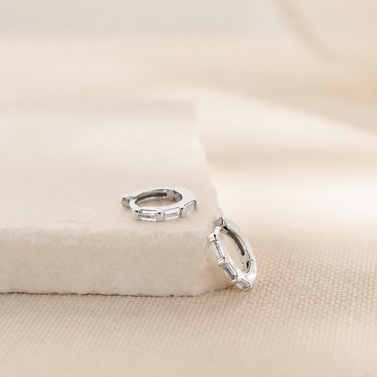 A pair of silver huggie hoop earrings with rectangular clear stones, displayed on a textured beige fabric surface with a soft beige block prop.