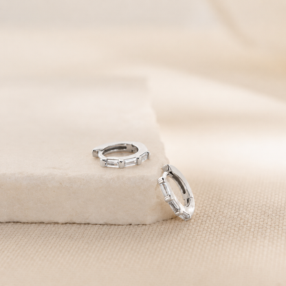 A pair of silver huggie hoop earrings with rectangular clear stones, displayed on a textured beige fabric surface with a soft beige block prop.