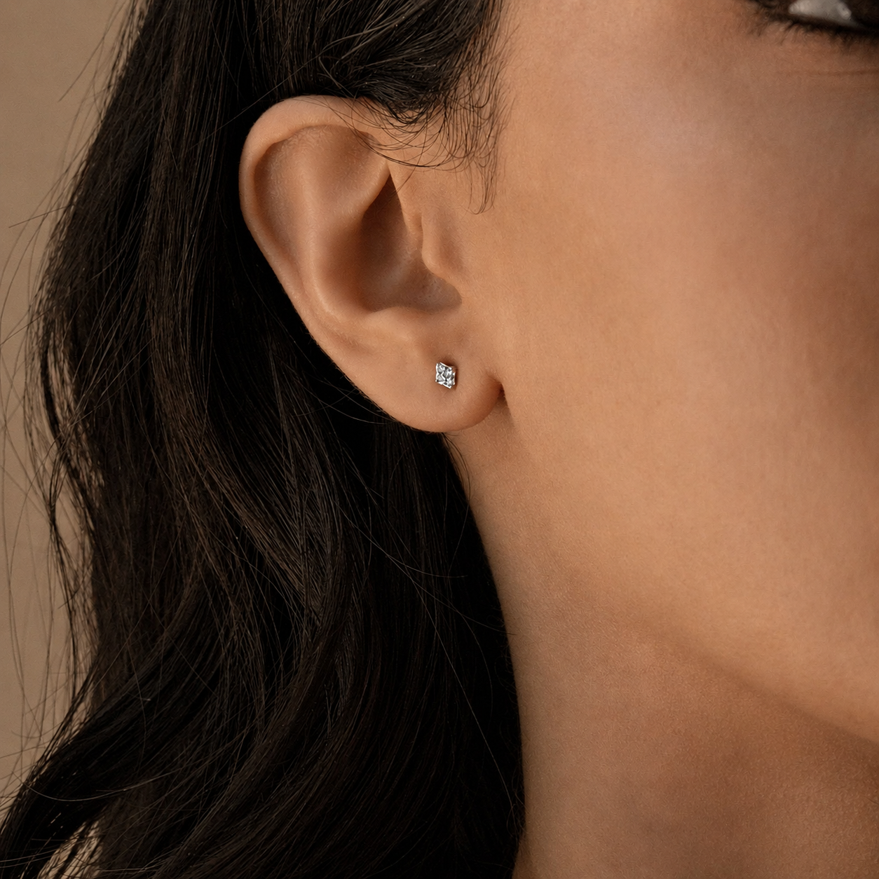 A close-up of a woman's ear wearing a small square stud earring with a sparkling clear gemstone.
