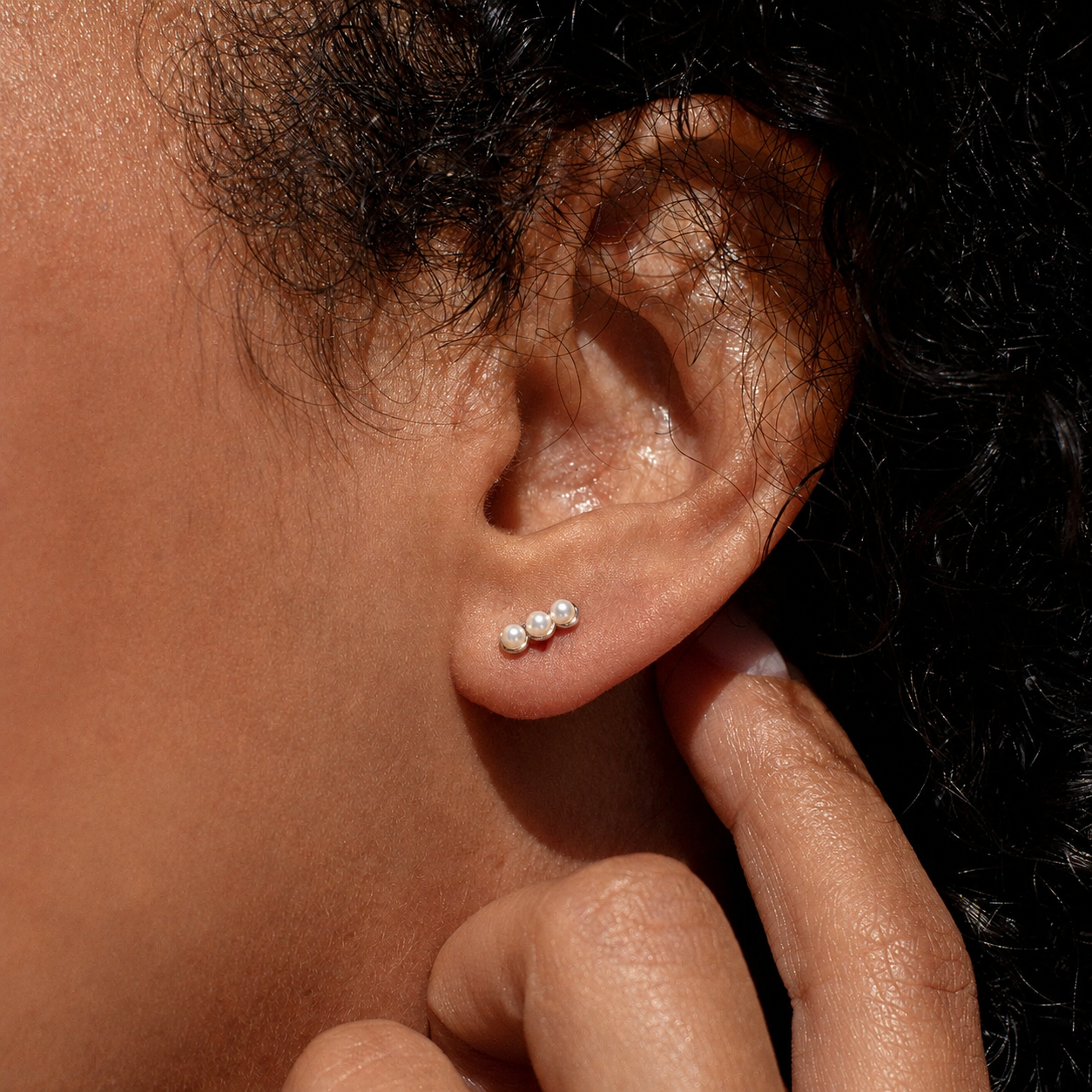 A close-up of a person's ear wearing a small cluster stud earring with three tiny white pearl-like beads on a dark background.