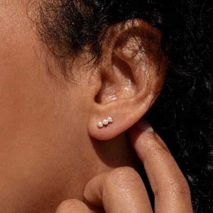 A close-up of a person's ear wearing a small cluster stud earring with three tiny white pearl-like beads on a dark background.