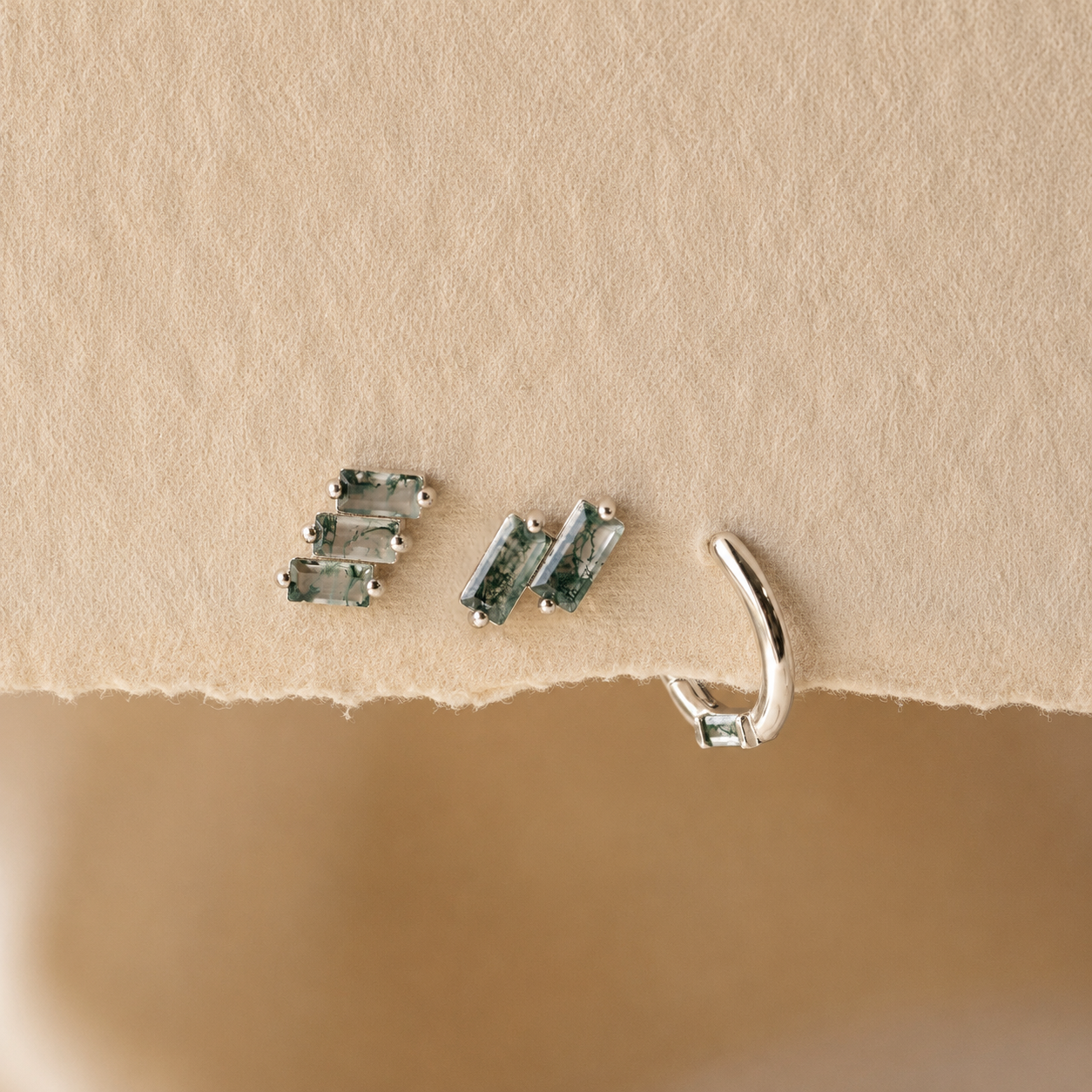 Three pairs of silver-toned ear cuff earrings with rectangular green-blue stones, displayed on a textured beige paper surface.