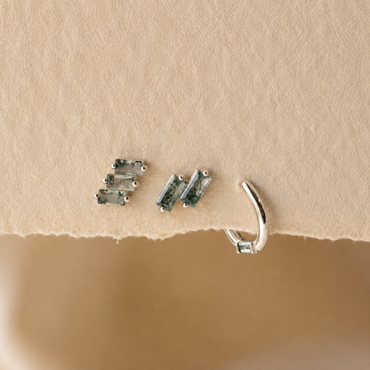 Three pairs of silver-toned ear cuff earrings with rectangular green-blue stones, displayed on a textured beige paper surface.