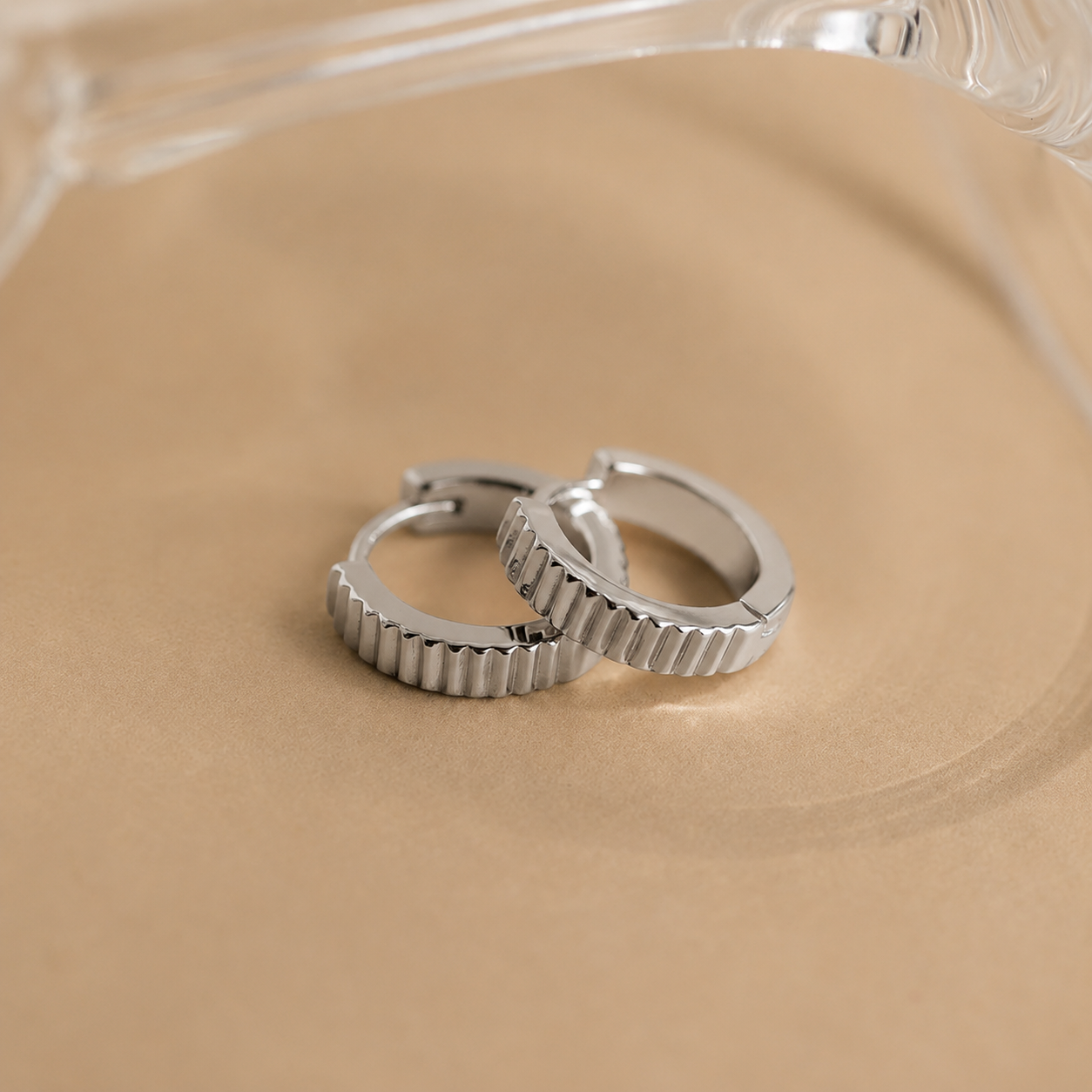 Two silver hoop earrings on a beige surface with a clear glass object in the background.