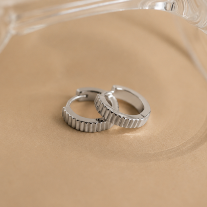 Two silver hoop earrings on a beige surface with a clear glass object in the background.