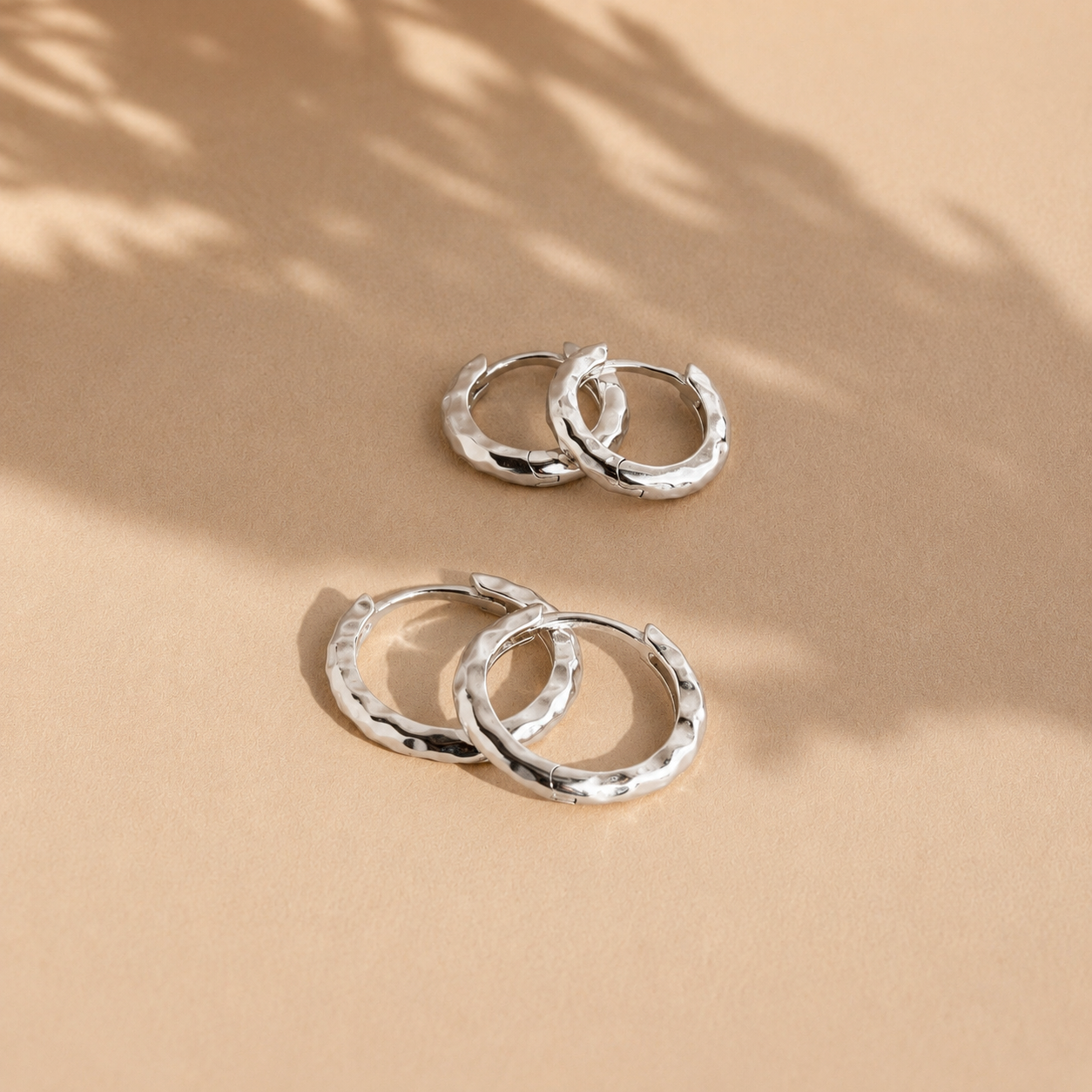 Pair of silver hoop earrings on a beige surface with natural light casting shadows.