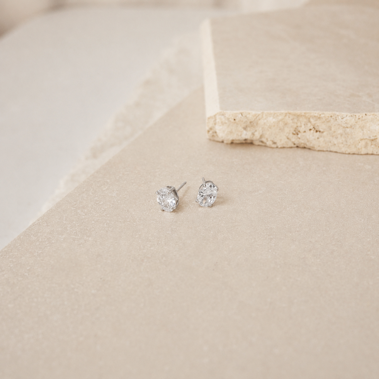 A pair of small silver stud earrings with clear gemstones, resting on a textured beige surface with a stone prop in the background.