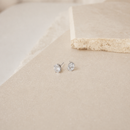 A pair of small silver stud earrings with clear gemstones, resting on a textured beige surface with a stone prop in the background.