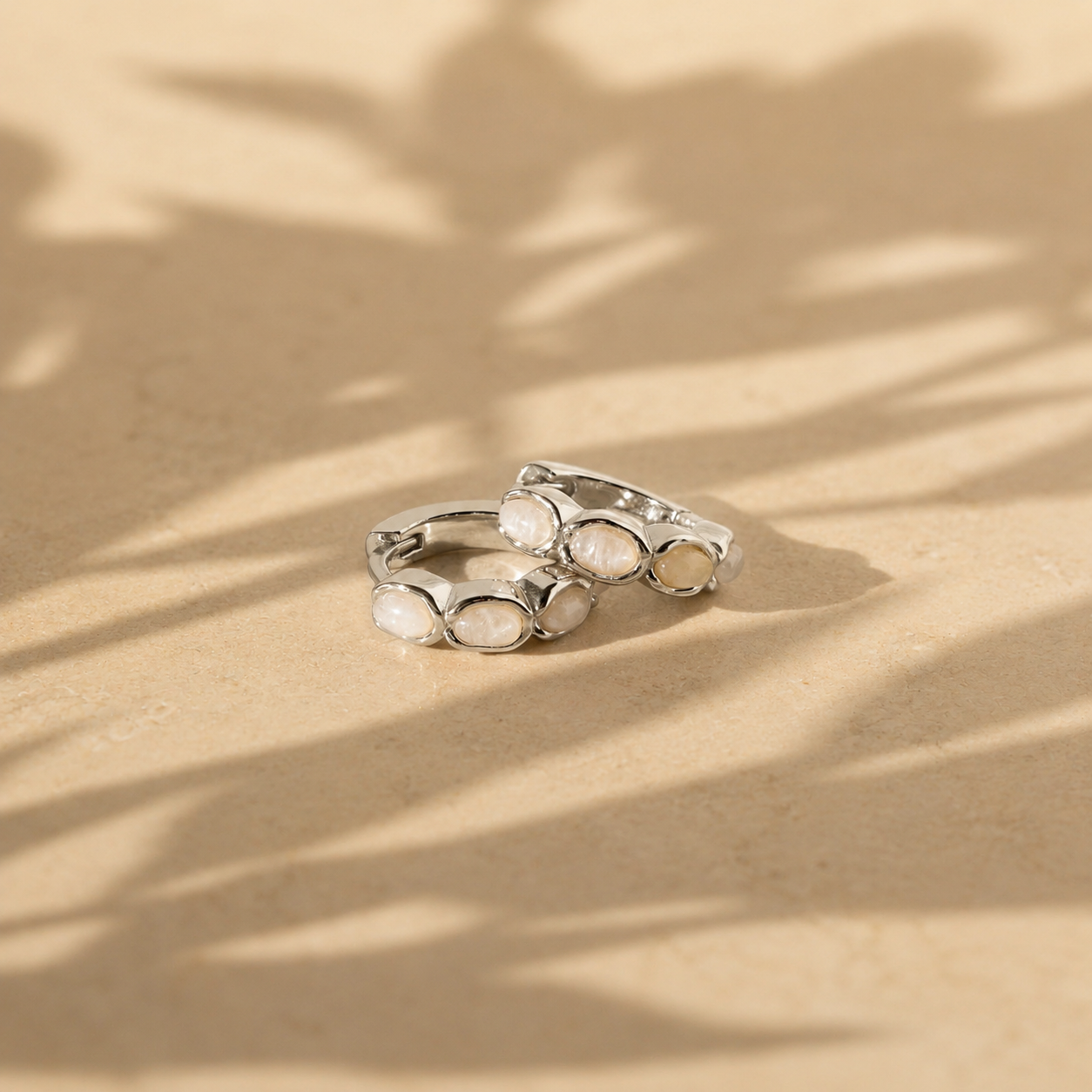 Silver hoop earrings with embedded stones on a beige surface with leaf shadows