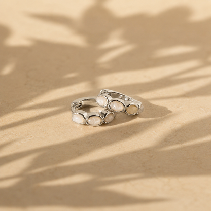 Silver hoop earrings with embedded stones on a beige surface with leaf shadows