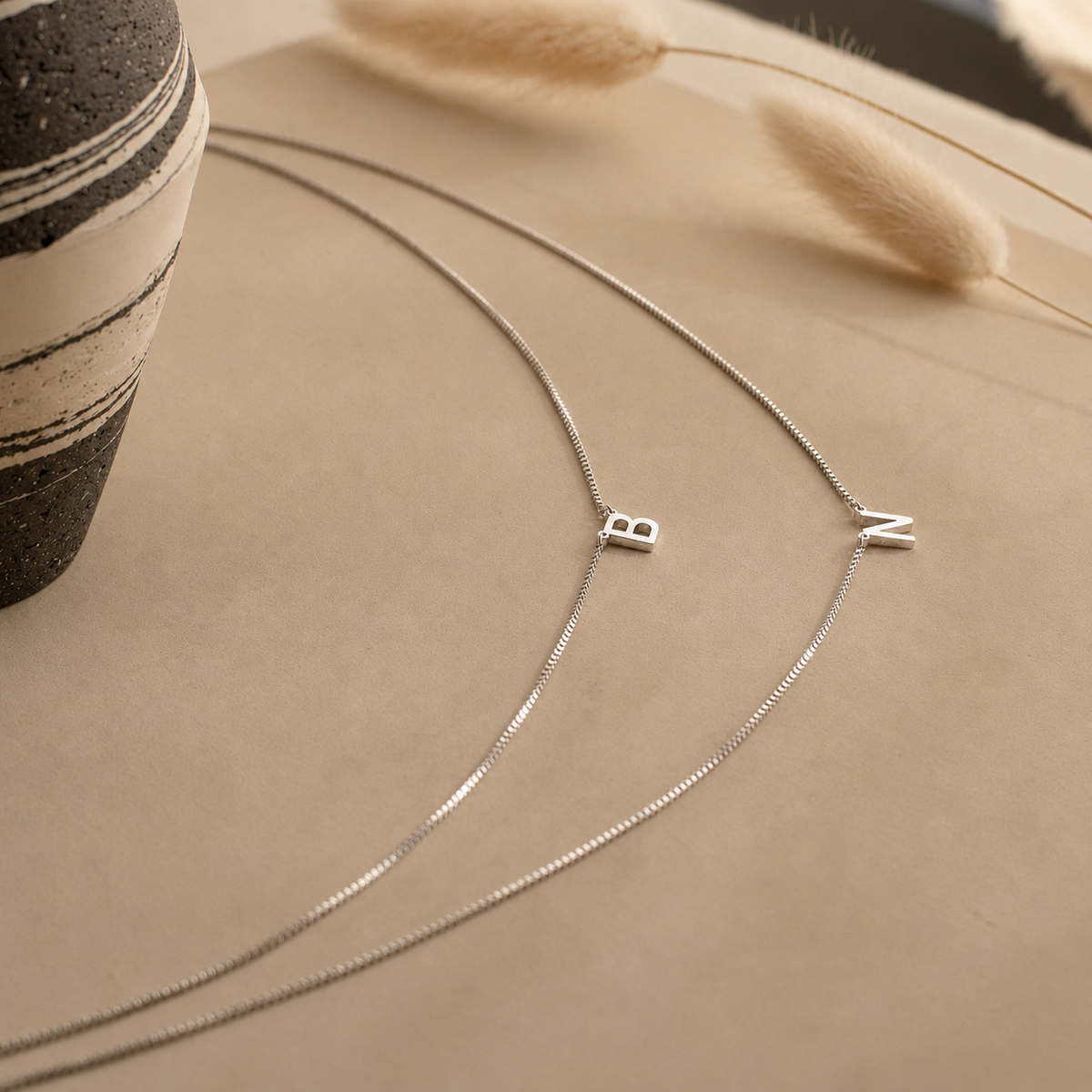 Silver necklace with 'BZ' charms on a beige surface with a vase and dried grasses.