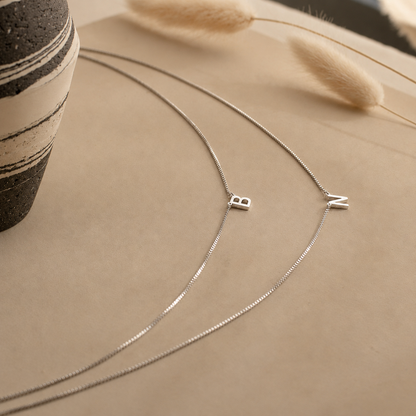 Silver necklace with 'BZ' charms on a beige surface with a vase and dried grasses.