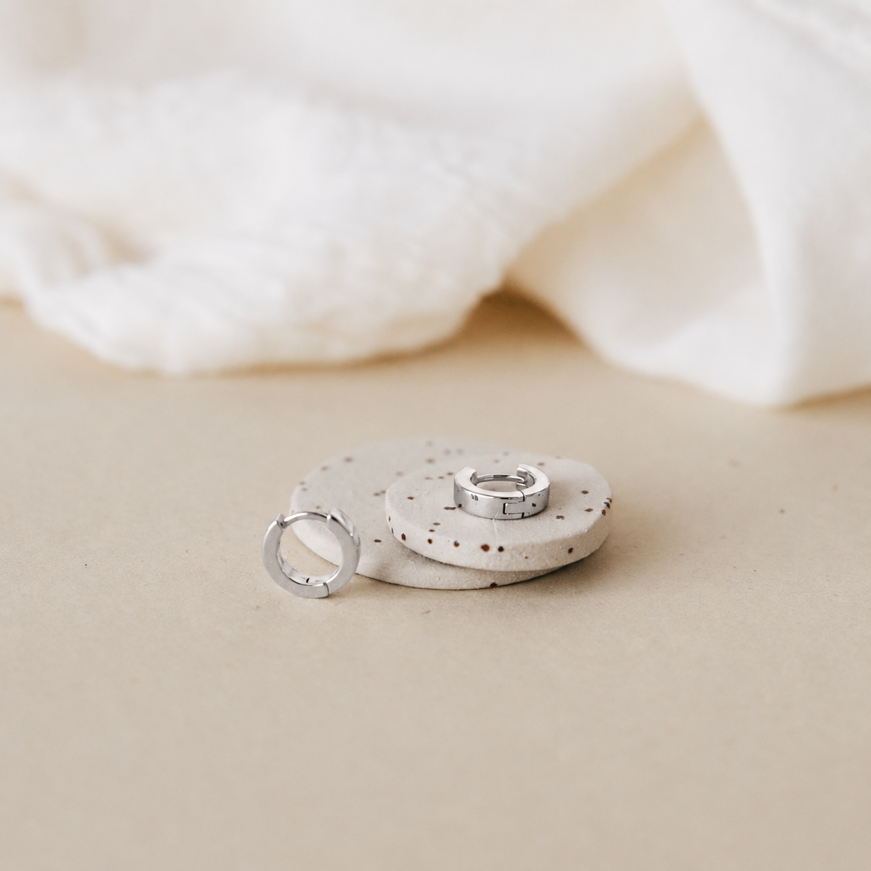 Two silver rings displayed on small round beige stones against a warm beige background with soft white fabric blurred in the background.