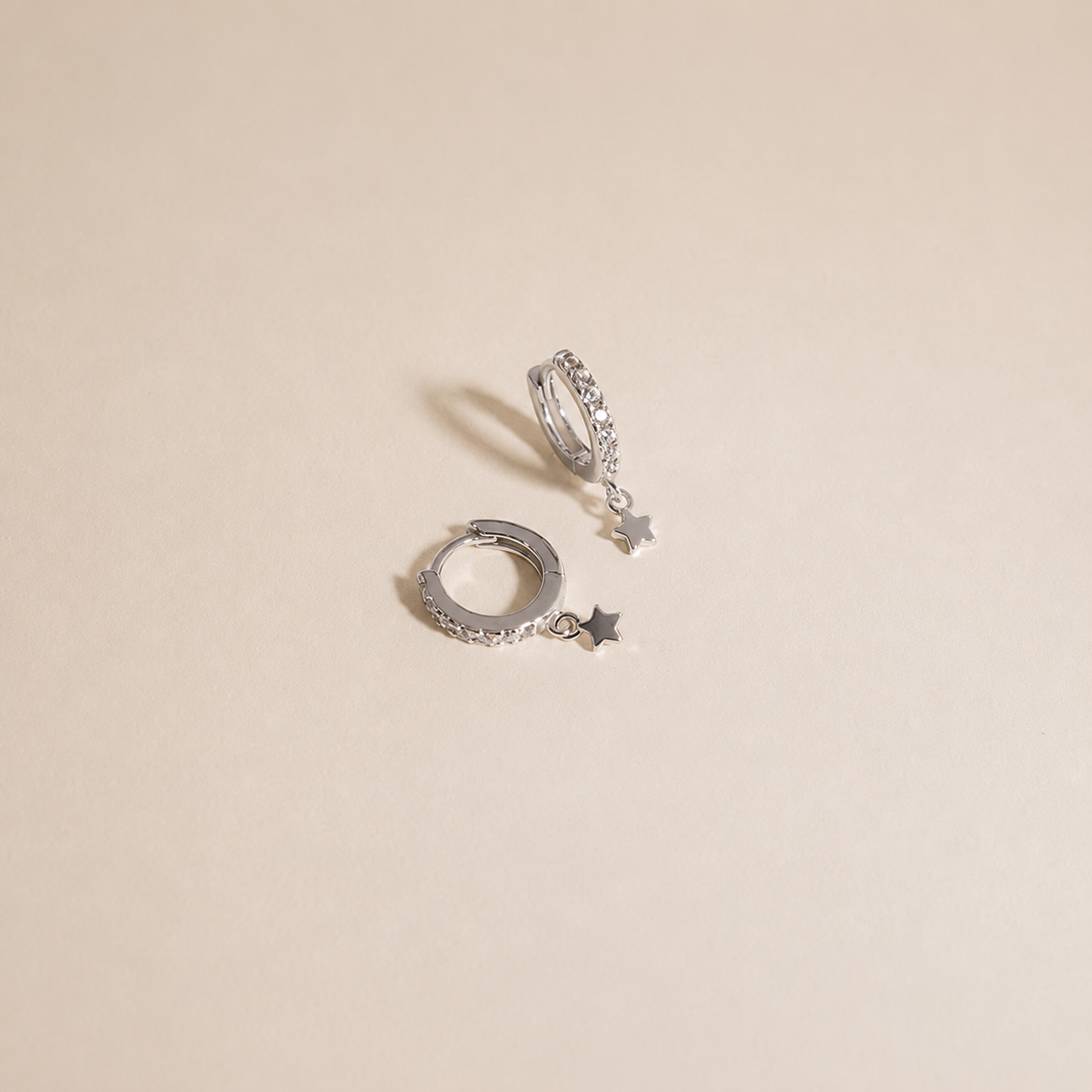A pair of silver huggie hoop earrings with small dangling star charms, displayed on a light beige background.