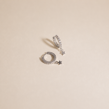 A pair of silver huggie hoop earrings with small dangling star charms, displayed on a light beige background.