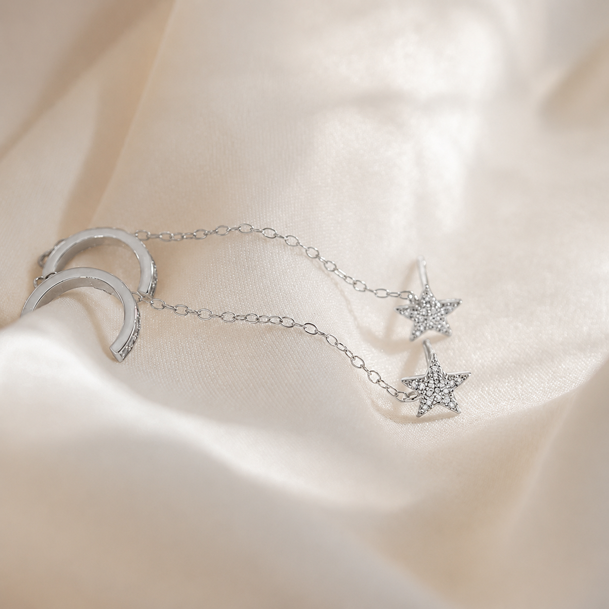 A silver crescent moon and star chain earring with clear rhinestone details, displayed on draped cream-colored satin fabric.
