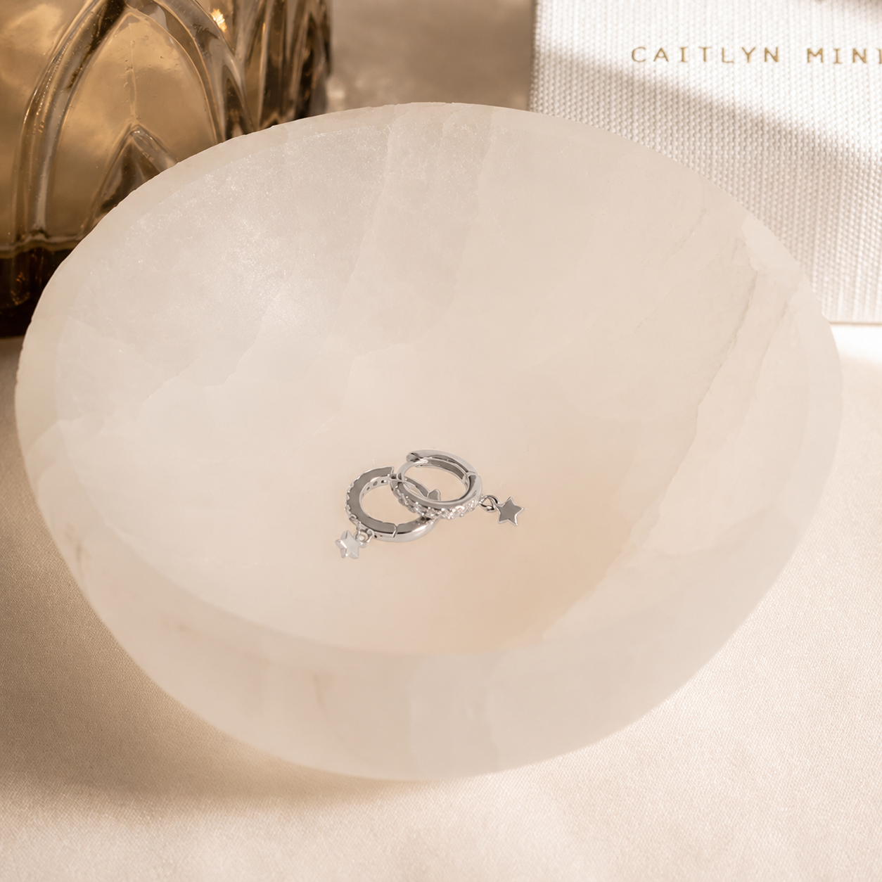 A pair of silver hoop earrings with small dangling star charms, displayed inside a translucent white bowl on a light beige surface.