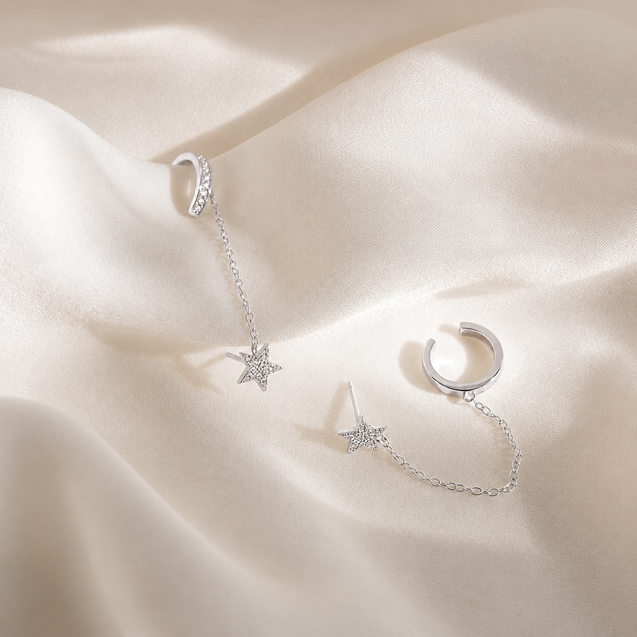 A pair of silver-toned huggie hoop earrings with delicate chain extensions and star-shaped charms, displayed on draped cream satin fabric.