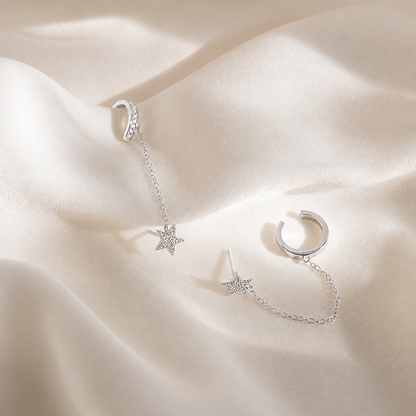 A pair of silver-toned huggie hoop earrings with delicate chain extensions and star-shaped charms, displayed on draped cream satin fabric.