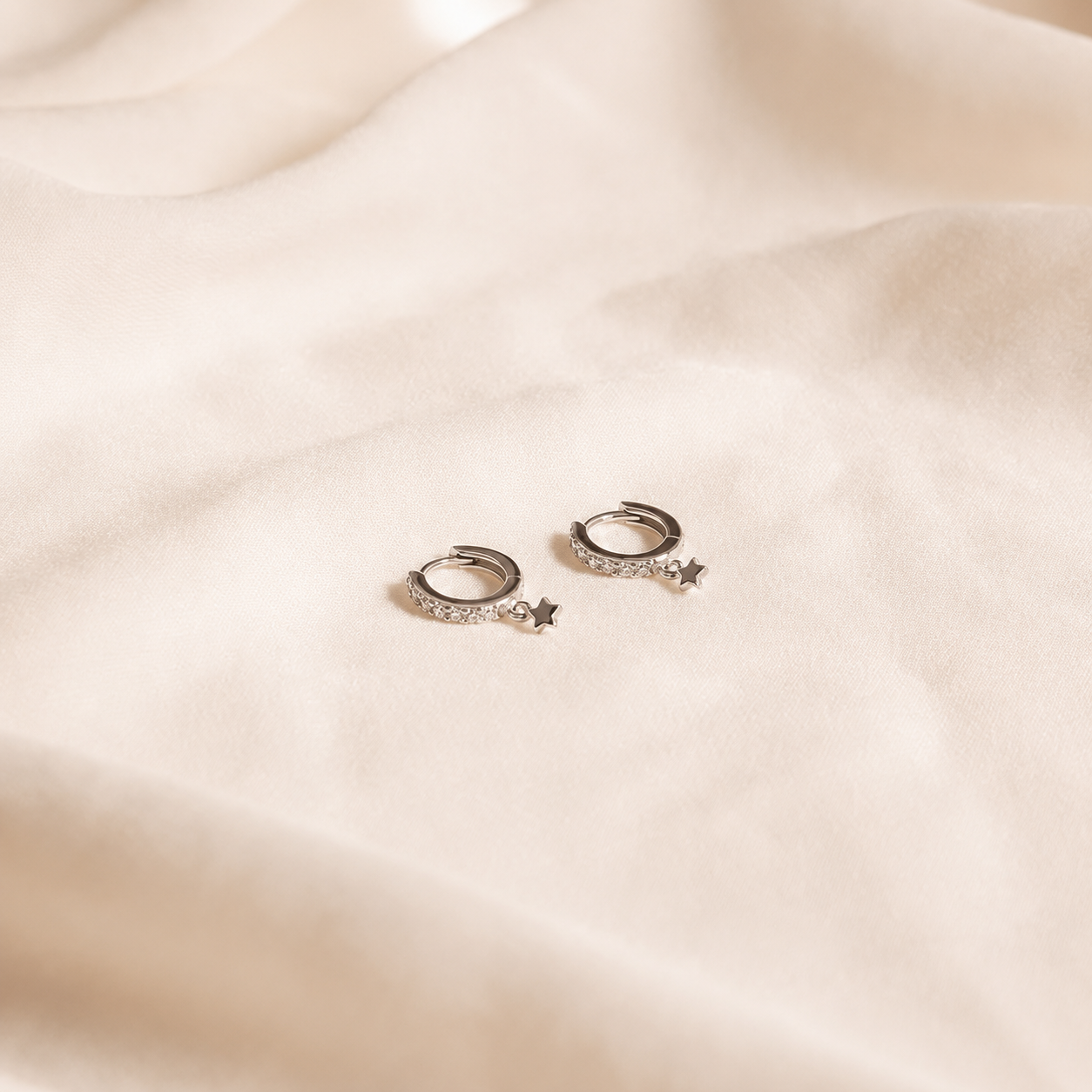 A pair of small silver hoop earrings with tiny clear stones and dangling star charms, placed on soft cream-colored fabric.