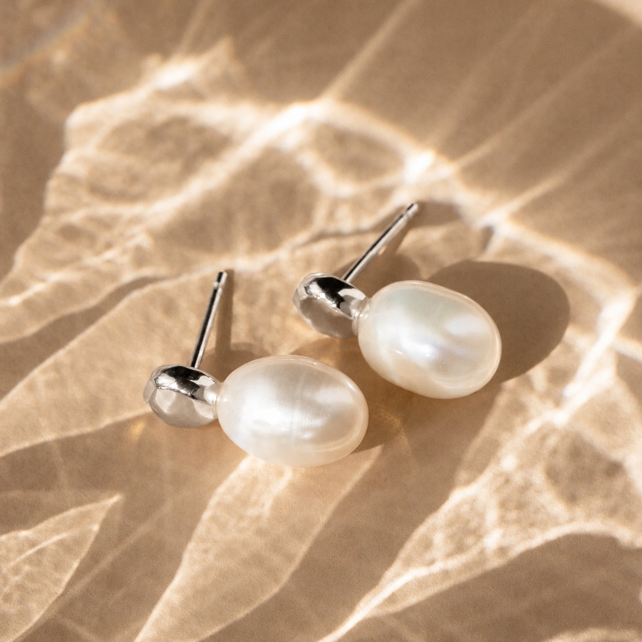 Pearl earrings on a textured beige surface with sunlight casting shadows