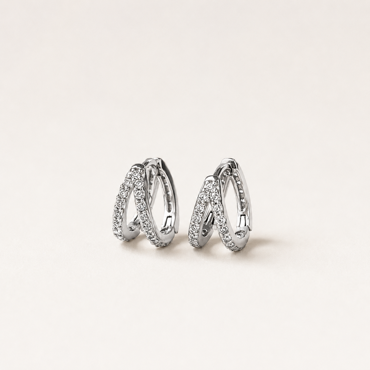 A pair of silver-toned huggie hoop earrings with pavé-set clear stones, displayed on a clean, light beige background.