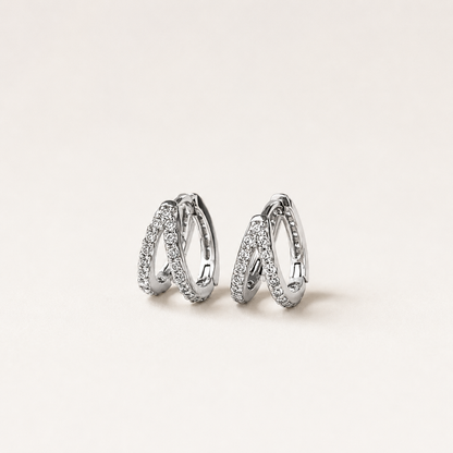 A pair of silver-toned huggie hoop earrings with pavé-set clear stones, displayed on a clean, light beige background.