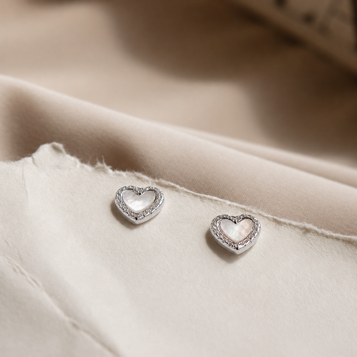 A pair of silver heart-shaped stud earrings with clear crystal accents, resting on a soft beige fabric background.